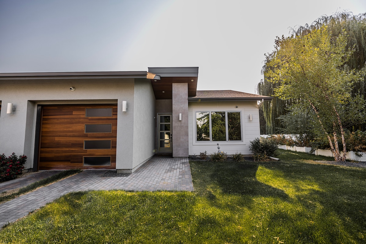 A modern guesthouse exterior is showcased, featuring a wooden garage door and well-maintained landscaping. The structure is complemented by large windows that allow natural light to brighten the interior. A neatly paved pathway leads to the entrance, flanked by manicured grass.