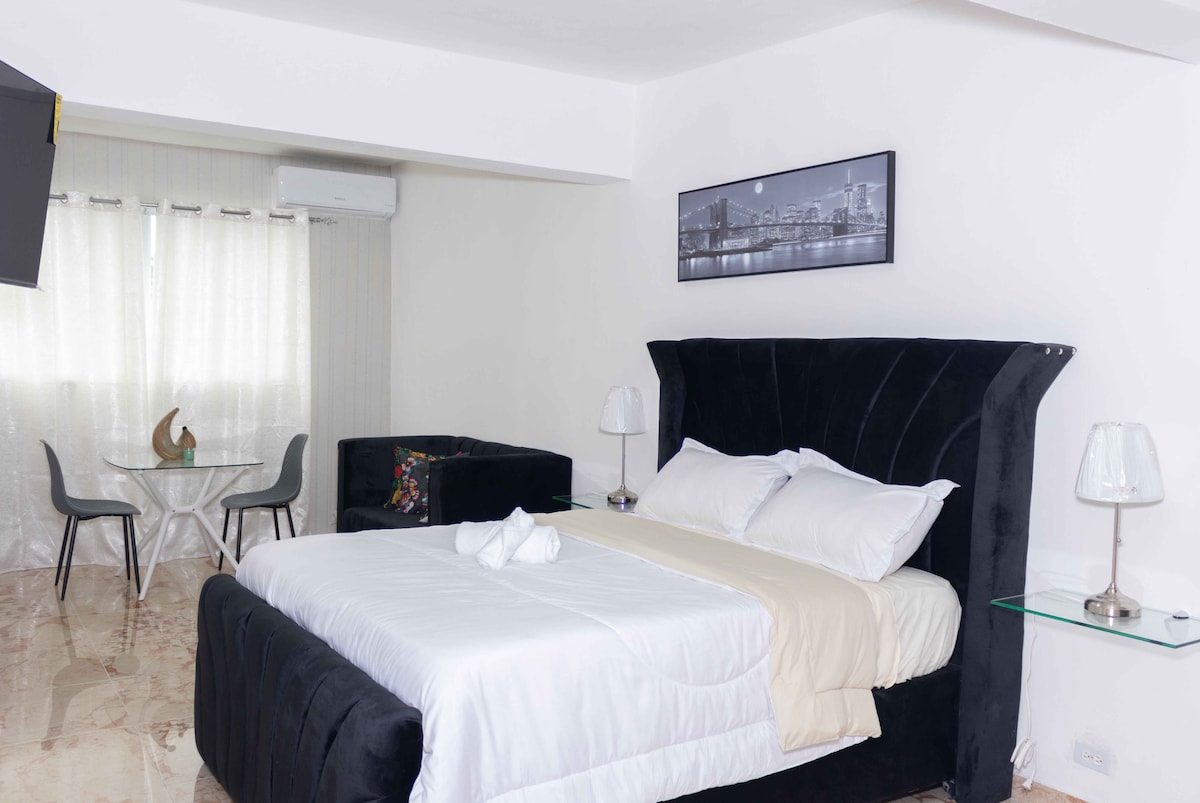 A spacious bedroom features a large bed with soft linens and a cozy decorative throw. A small dining area with a round table and chairs is present, along with a comfortable seating area. Light filters through sheer curtains, enhancing the room's bright ambiance.