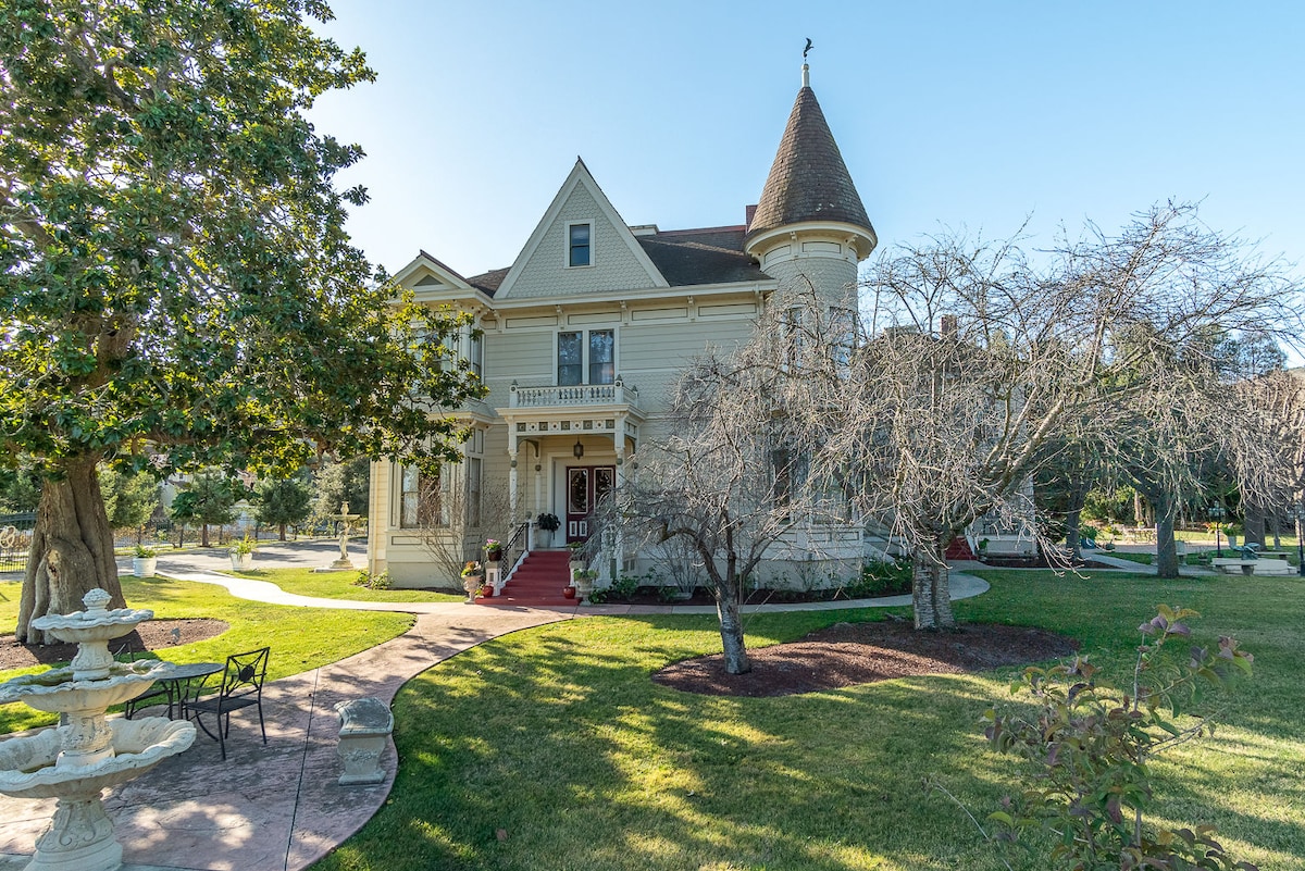 A historic mansion with an elegant design features a turret and a wraparound porch. The property is set on a landscaped lawn with mature trees and a decorative fountain in the foreground, inviting tranquility and charm.
