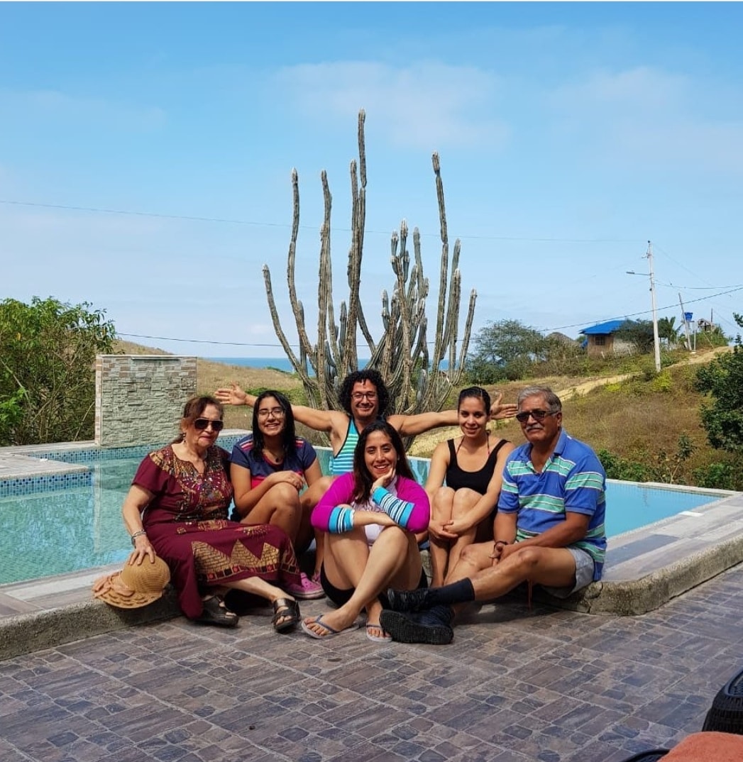A group of individuals is seated by a serene pool, with a backdrop of natural landscaping. The pool reflects the sky, and tall cacti are visible in the distance. The setting suggests a relaxed atmosphere ideal for gatherings.