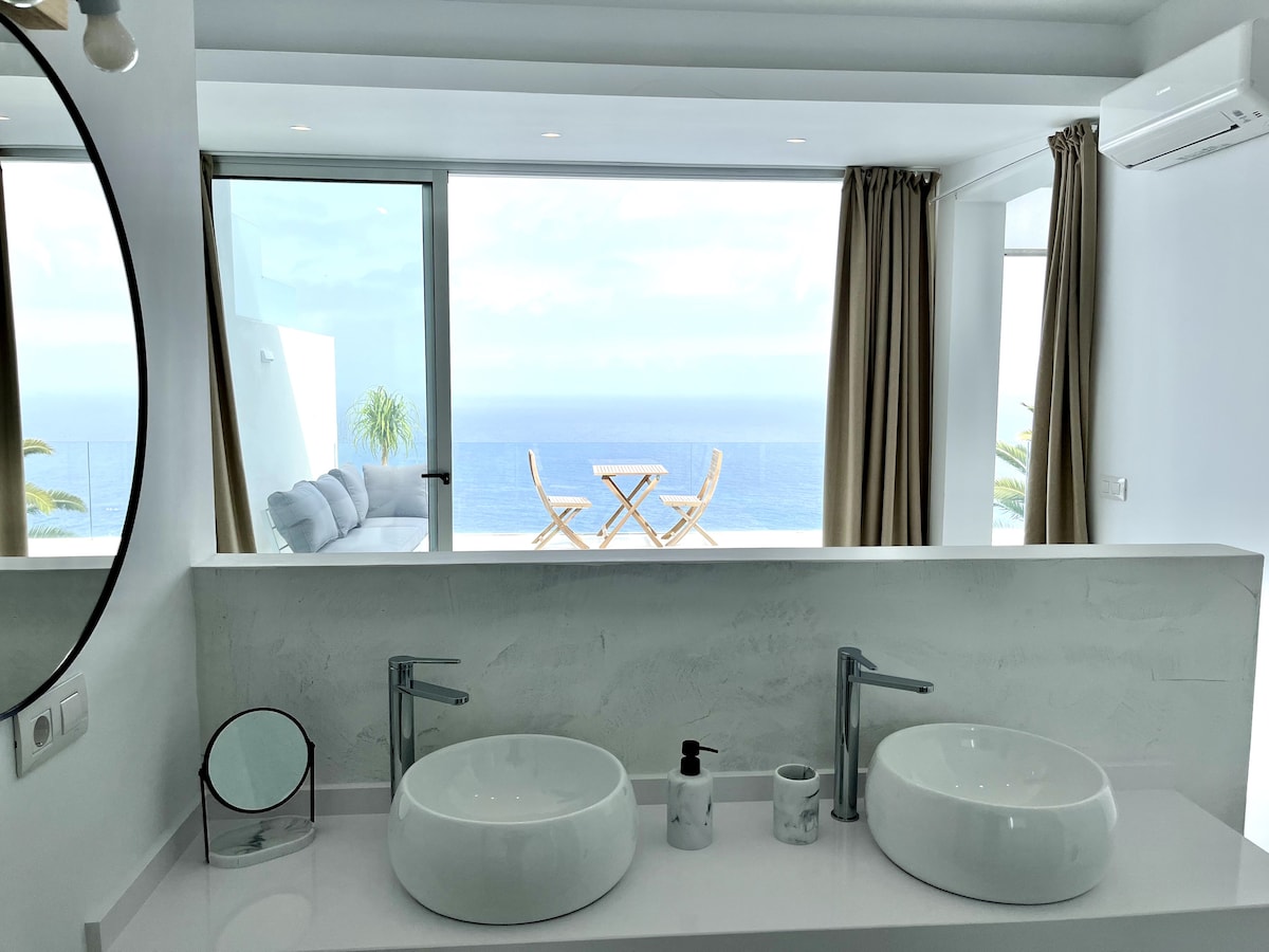 A modern bathroom features dual sinks with sleek faucets, positioned against a white countertop. A large circular mirror reflects the ample natural light flooding in from the expansive sliding glass doors, which offer a stunning view of the ocean and outdoor seating area.