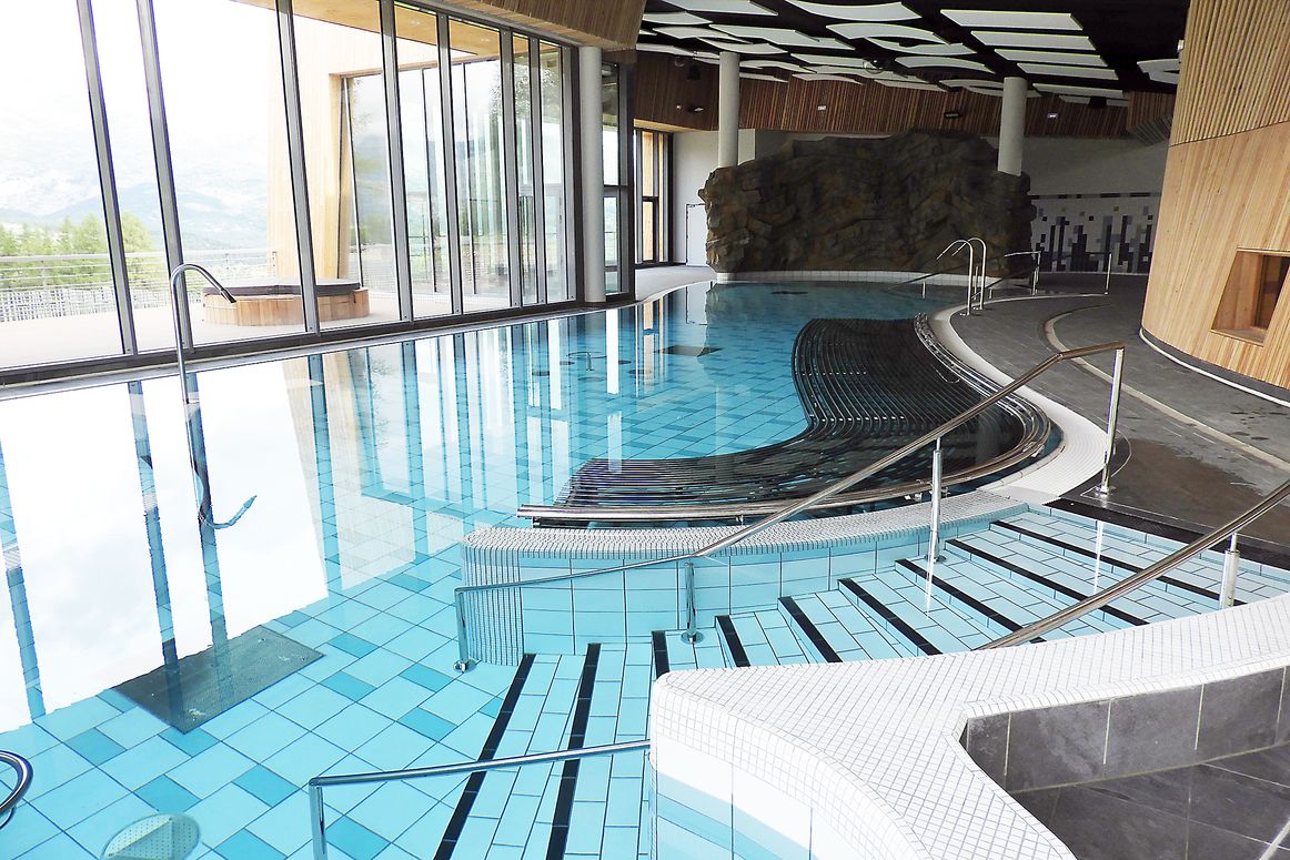 A spacious indoor pool area is highlighted, featuring a blend of turquoise tiled flooring and sleek black accents. Large windows provide an abundance of natural light and views of the surroundings, while a rocky feature adds a natural element to the contemporary design.