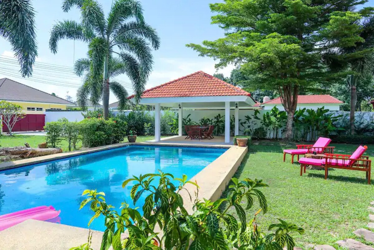 A private swimming pool is surrounded by a well-maintained garden featuring tropical plants. A dining sala with a red-tiled roof stands nearby, with seating arrangements visible. Sun loungers are placed on the grass, inviting relaxation in the serene outdoor space.