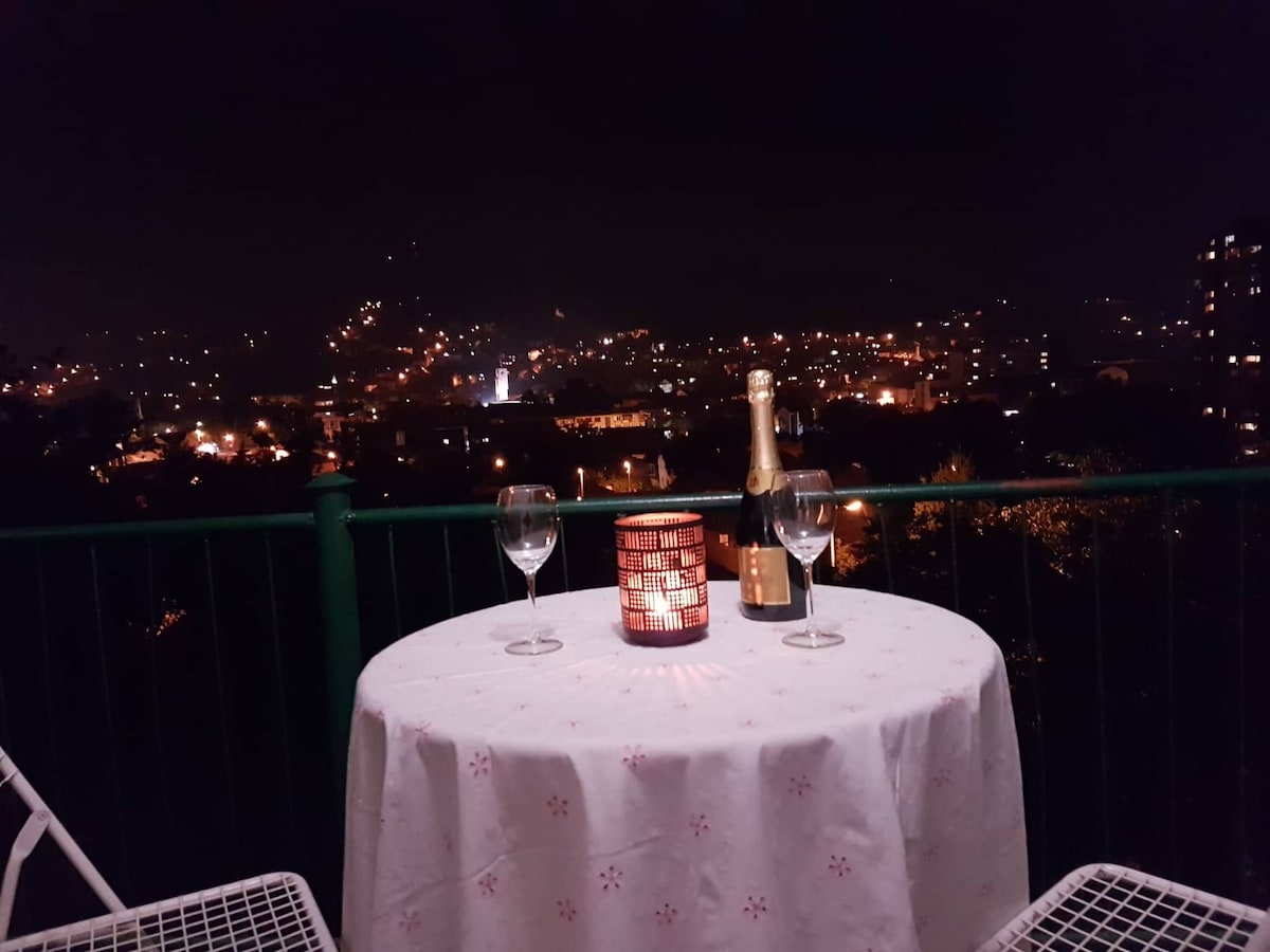 A small outdoor table is set for two, featuring a white tablecloth and a lit candle. Two glasses sit alongside an opened bottle of champagne. The view captures a twinkling night skyline, with the lights of the city creating a serene atmosphere.