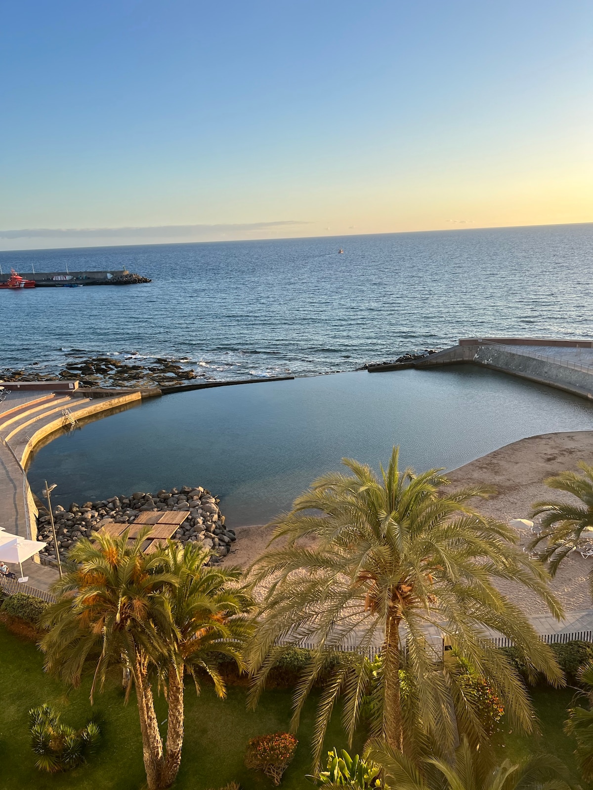 A serene view is presented, showcasing calm water meeting the horizon. Palm trees gently frame the foreground, while a pedestrian walkway leads toward the water's edge. The soft hues of sunset illuminate the scene, reflecting off the tranquil surface.