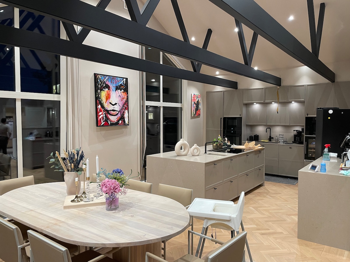 A modern kitchen features high ceilings with exposed beams, enhancing the spacious feel. A round dining table is set with a vase of flowers, while the kitchen island displays various utensils. Sleek cabinetry and high-end appliances are visible, providing a functional cooking space.