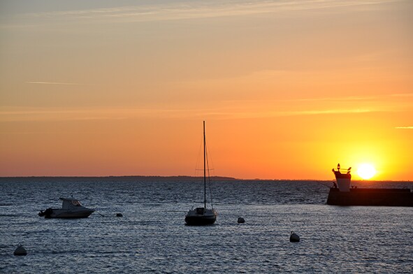 The image captures a serene sunset over the water, with two boats gently bobbing on the waves. The sun, partially submerged, casts a warm glow across the horizon, creating a tranquil atmosphere. Silhouetted against the sunset, a beacon stands by the shore.