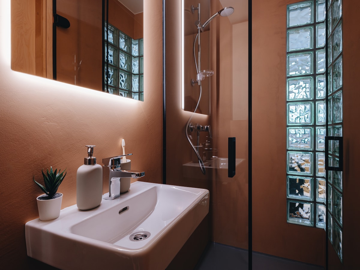 The bathroom showcases a minimalist design with a sleek sink and modern fixtures. Soft lighting illuminates the space, highlighting the glass block shower area. A small potted plant adds a touch of greenery, complementing the warm-toned walls.