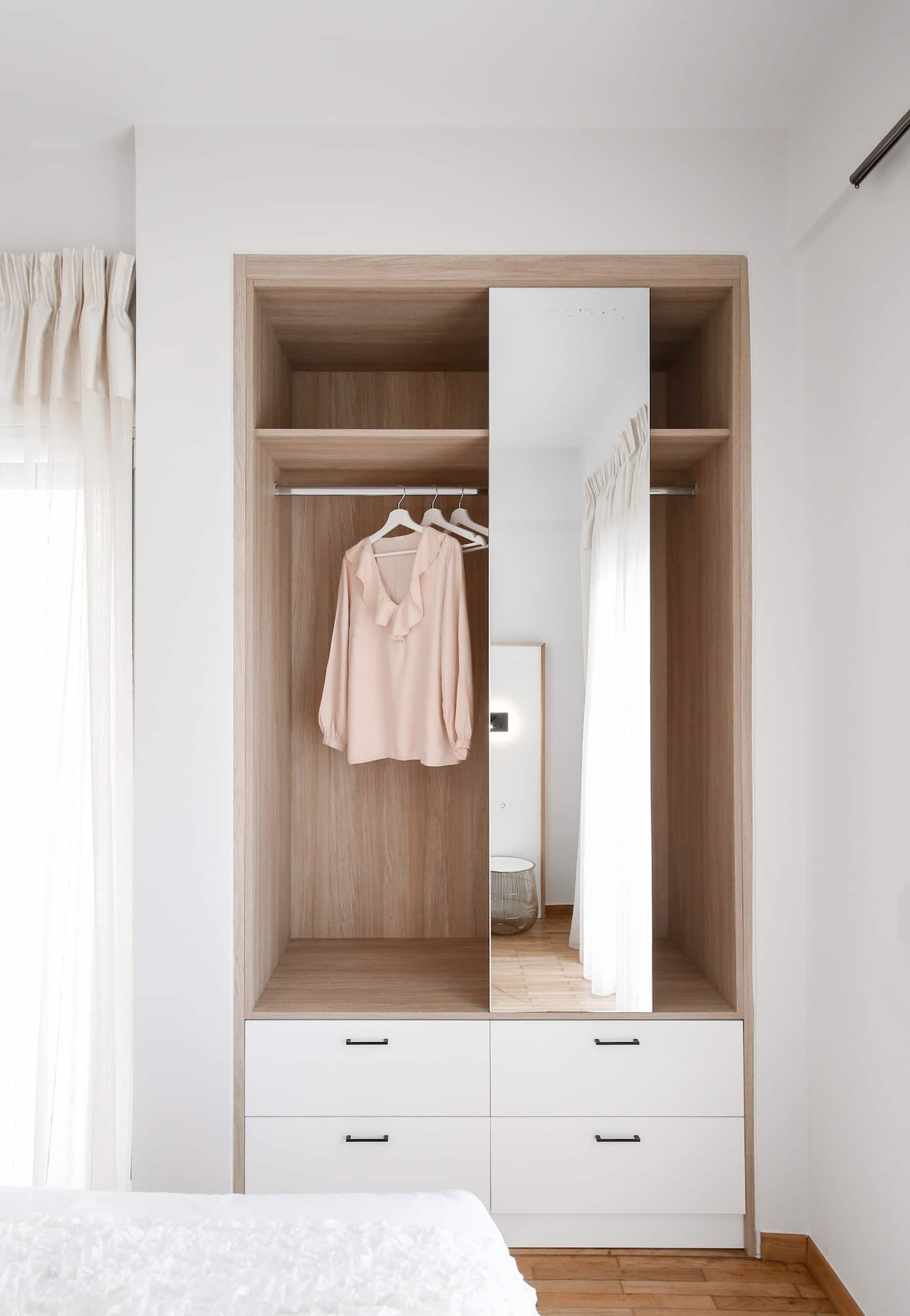 A modern wardrobe features a wooden finish with a large mirror front. Hangars display a soft pink blouse, while below, drawers offer additional storage. Natural light filters through sheer curtains, creating a bright and airy feel in the space.
