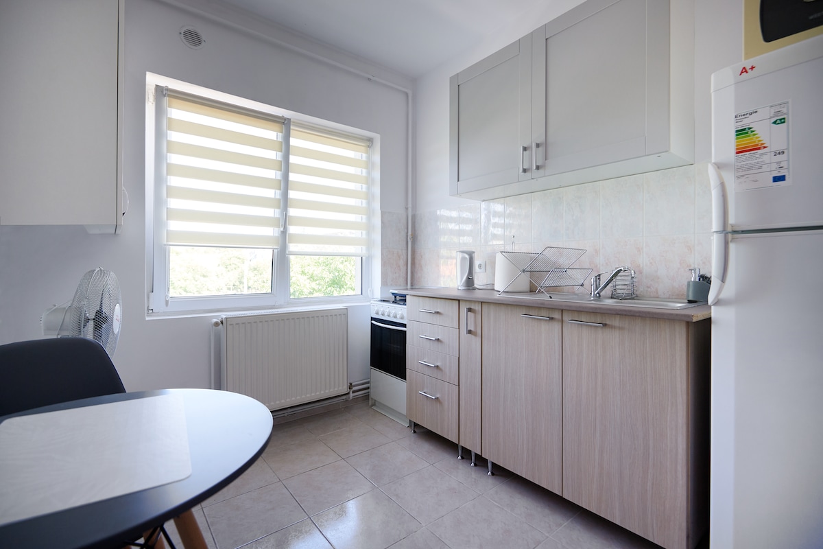 A functional kitchen is equipped with modern cabinetry and a combination of appliances, including a stove and refrigerator. Natural light enters through a large window with window coverings, enhancing visibility. A small dining table and a fan are positioned, contributing to a practical layout.