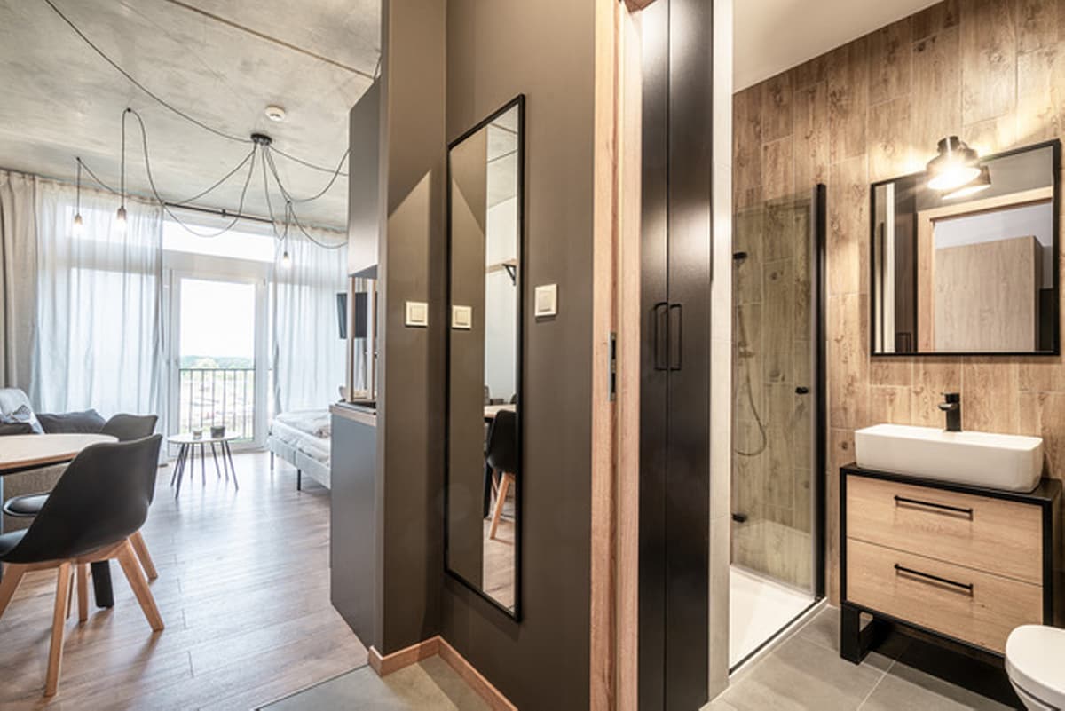 An open layout reveals a modern living space with a comfortable seating area and dining table. A large mirror reflects natural light, while a stylish bathroom features a walk-in shower and contemporary sink. Neutral tones and wood accents create a cohesive aesthetic.