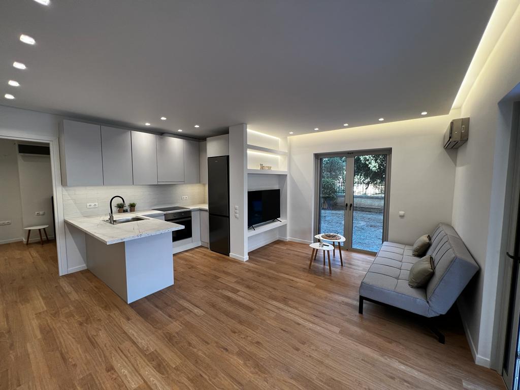 A modern open-plan living area features a dual-purpose sofa bed and a sleek kitchen counter with a minimalist design. Soft overhead lighting enhances the wooden flooring, while glass doors lead to a view of the garden. A wall-mounted television is integrated into the space.