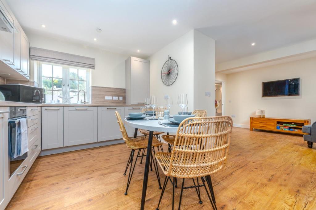 A modern kitchen and dining space features a circular table set with tableware. Four stylish woven chairs surround the table. The kitchen includes sleek cabinetry and high-end appliances, while large windows allow natural light to fill the area, enhancing the warm wood flooring.