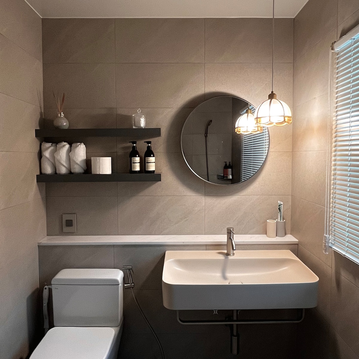 A modern bathroom is featured, consisting of a sleek sink with a minimalist design, and a round mirror above it. Grey tiled walls are complemented by dark shelving displaying toiletries. Soft lighting is provided by a pendant fixture, and natural light filters through a window adorned with blinds.