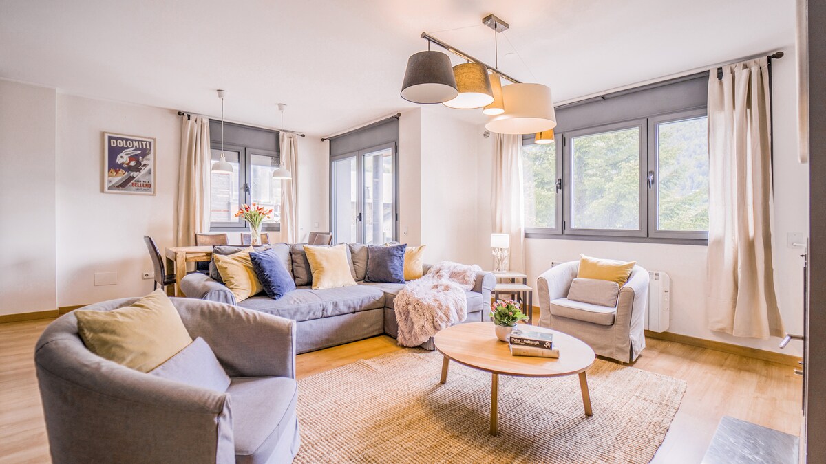 The living area features a light and airy design, with a spacious sofa arranged around a round coffee table. Two armchairs provide additional seating, while natural light filters through multiple windows adorned with soft curtains.