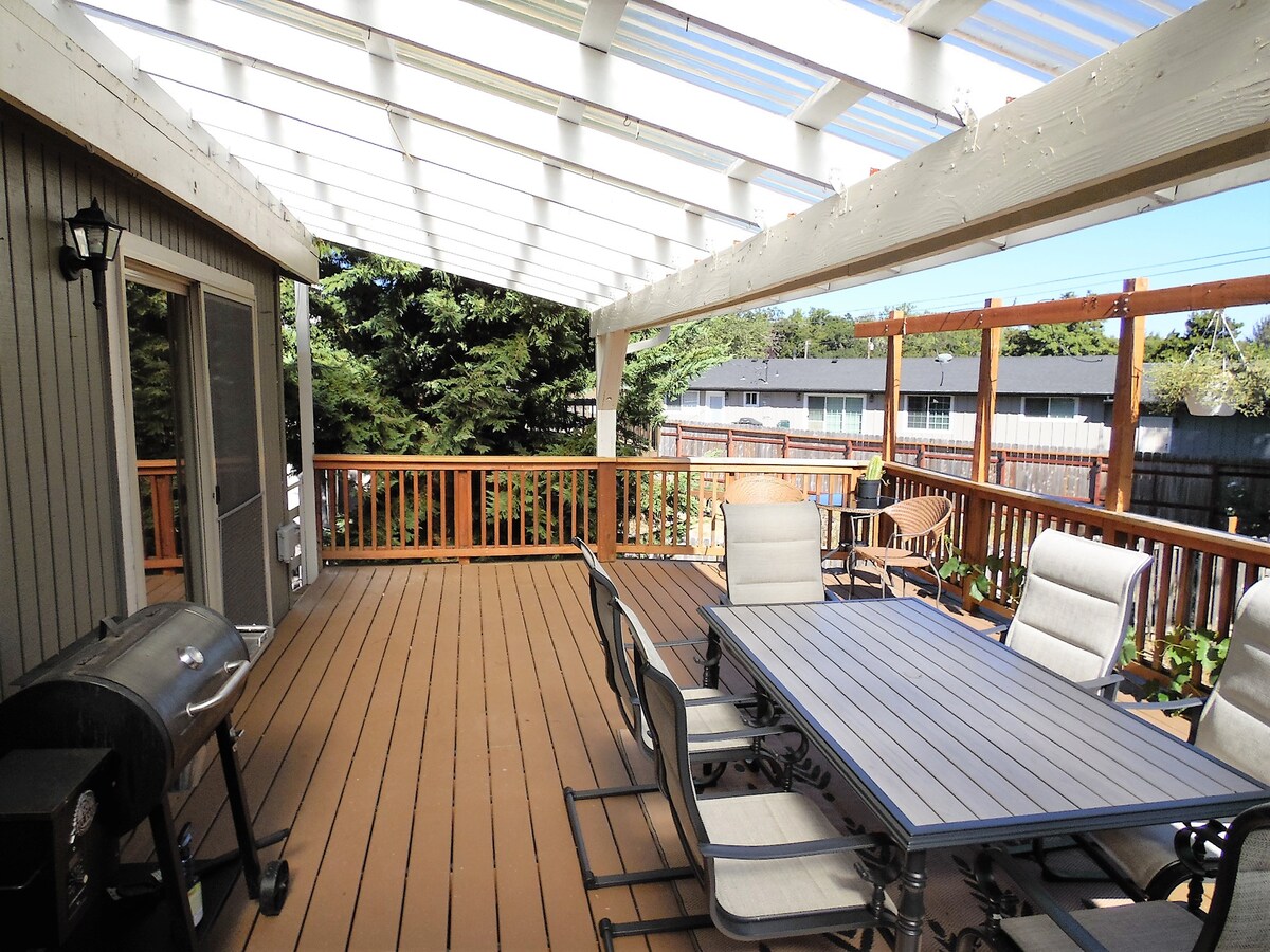 A spacious outdoor deck is furnished with a metal dining table and several chairs, creating an inviting dining area. A gas grill is positioned nearby, and the space is partially shaded by a translucent roof, providing comfort for outdoor gatherings.