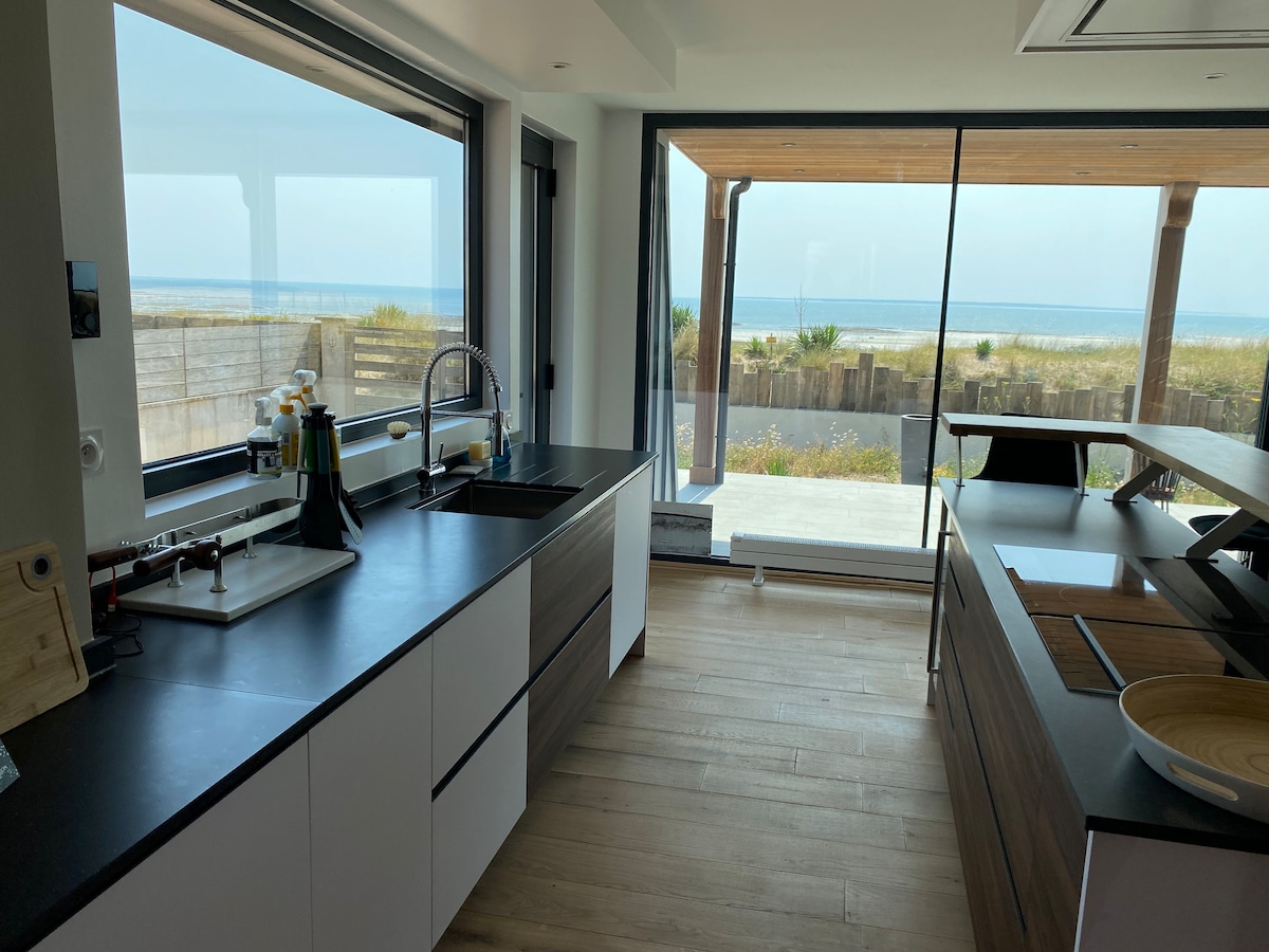 A modern kitchen area is displayed with sleek cabinetry and a dark countertop. Large windows provide a view of the beach and dunes outside. The space is filled with natural light, emphasizing a minimalist design. An island counter is visible, enhancing the open atmosphere.
