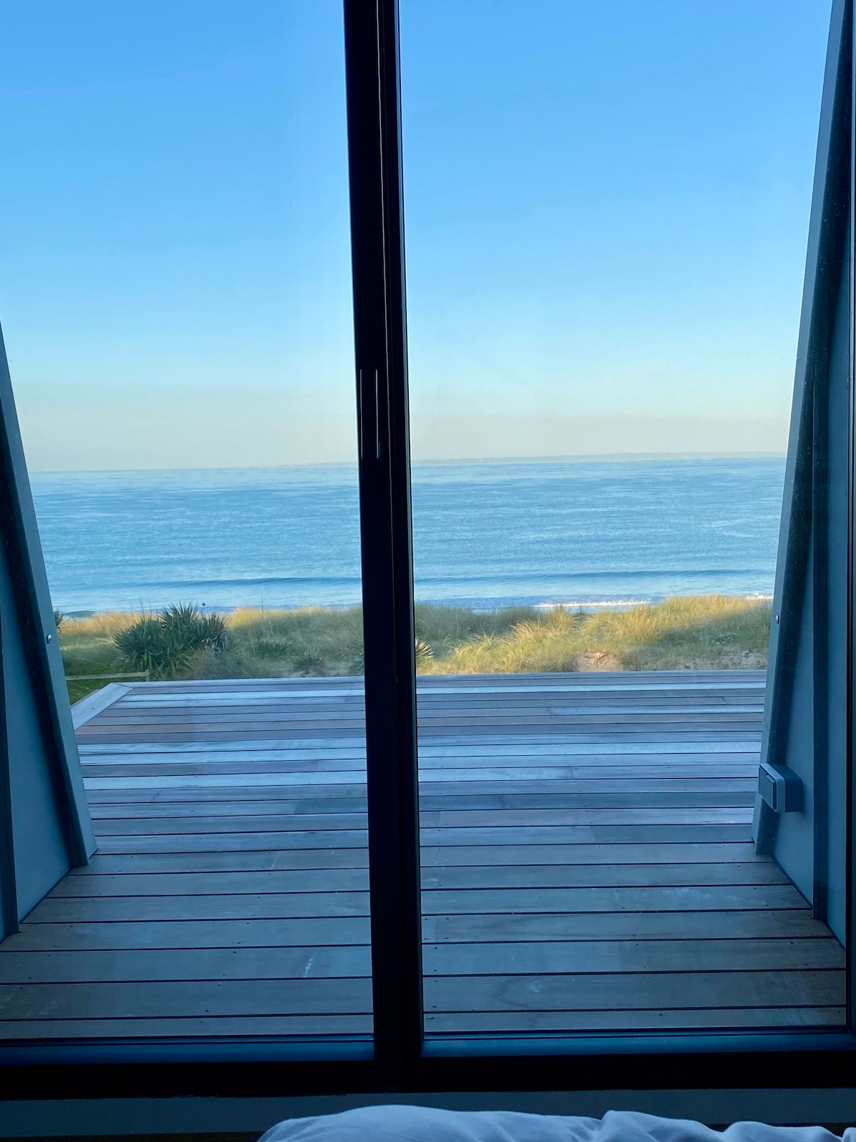 A panoramic view showcases the serene blue ocean, visible through expansive glass doors that open onto a wooden deck. The horizon meets soft sandy dunes, creating a tranquil coastal atmosphere.