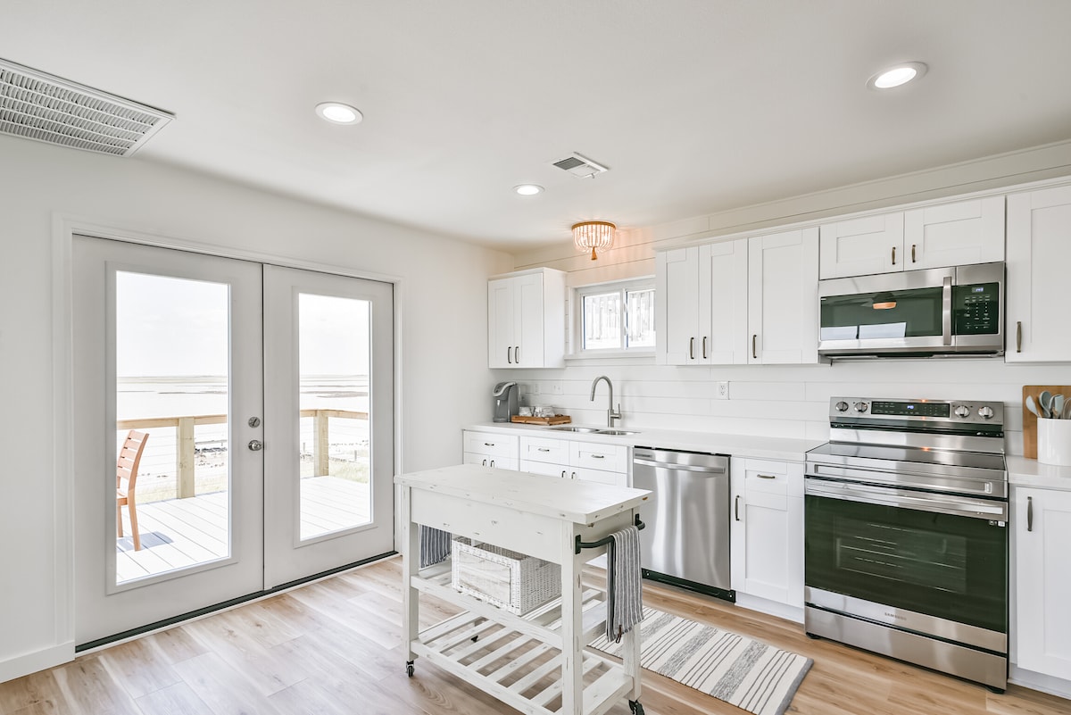 A modern kitchen features sleek white cabinets and stainless steel appliances, including an oven and dishwasher. A kitchen island with a rolling cart is positioned in the center. Two French doors open to a scenic outdoor deck, allowing natural light to fill the space.