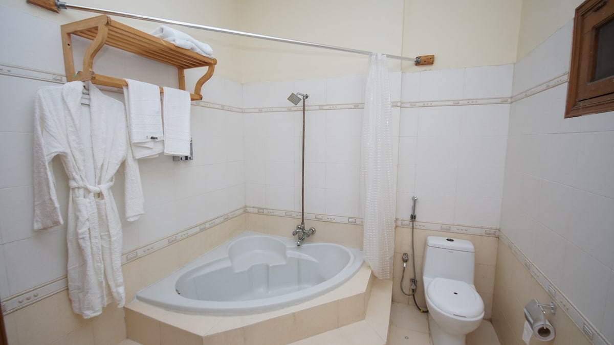 A bathroom is presented with a curved bathtub, accompanied by a showerhead and a white curtain. Towels are neatly arranged on a wooden shelf, and a plush bathrobe is hung beside the towels. A modern toilet is located adjacent to the bathtub, contributing to the functional layout.