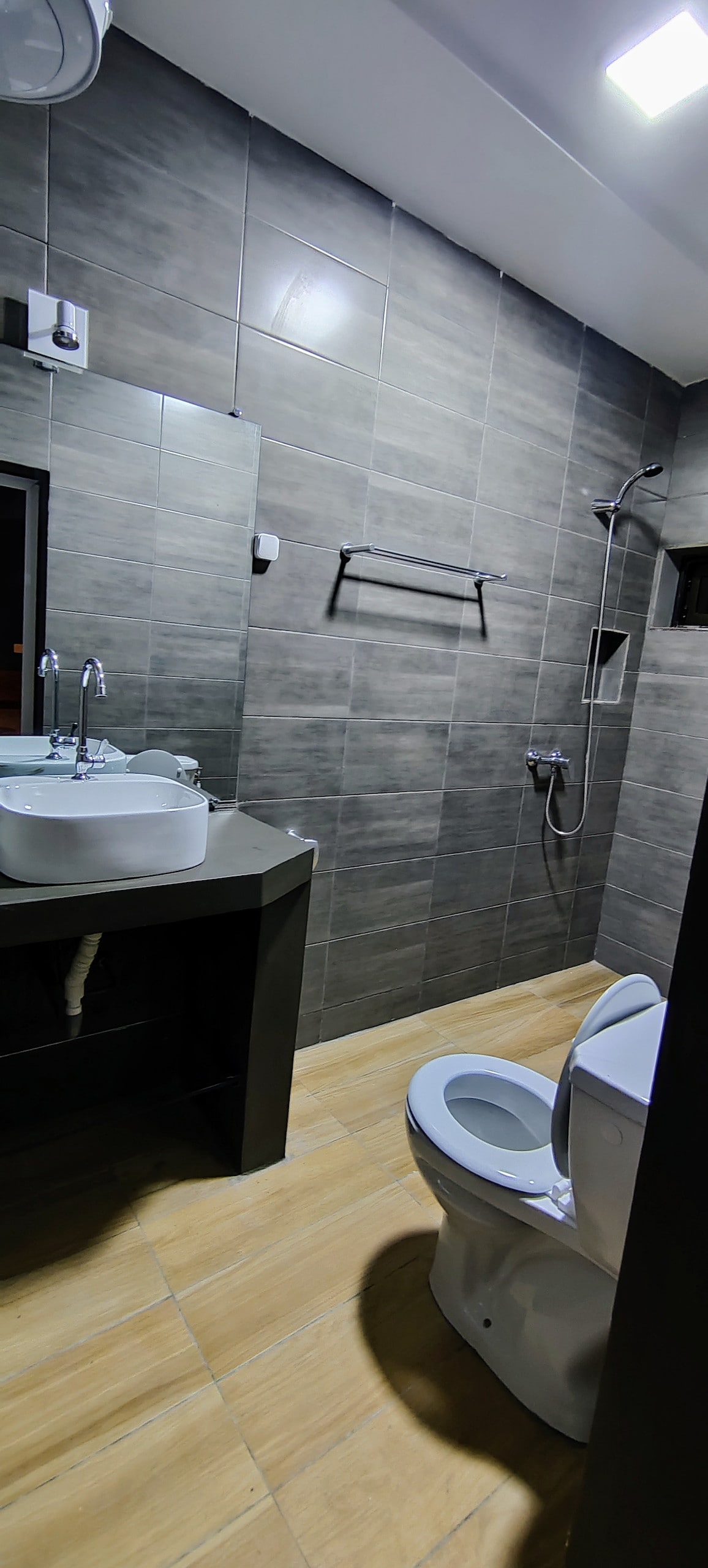 A modern bathroom is showcased with grey tiled walls and warm wooden flooring. A circular sink rests on a dark vanity, while a toilet is positioned adjacent. A shower equipped with a glass enclosure is visible, featuring a handheld showerhead and wall-mounted controls.