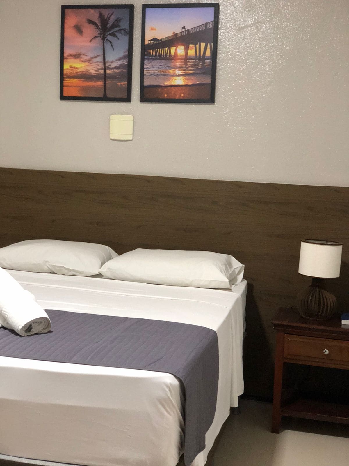 A standard bedroom is featured, showcasing a spacious bed with two neatly arranged pillows. A gray blanket is positioned across the bed. A bedside table with a lamp sits next to the bed, while two framed images of a pier and sunset adorn the wall.