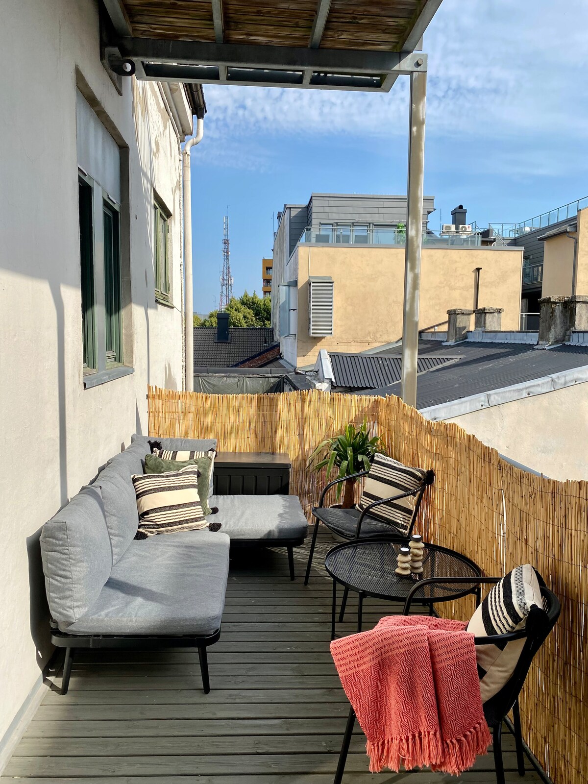 A balcony area is presented with a comfortable seating arrangement featuring gray couches and striped pillows, complemented by two small tables. A warm throw is draped over one chair. The surrounding bamboo fencing provides privacy, and distant city buildings are visible against a blue sky.