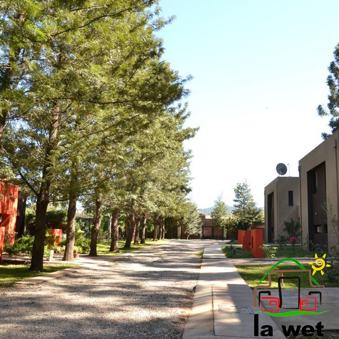 A gravel pathway is flanked by tall trees and colorful buildings, creating a serene and natural setting. The pathway leads through a landscaped area, showcasing a blend of greenery and architectural elements, promoting a peaceful atmosphere.