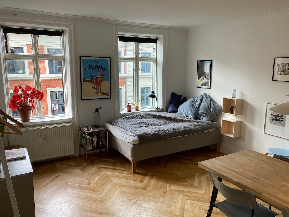 A light-filled room features a spacious bed with a gray blanket, complemented by decorative wall art and wooden accents. Two large windows provide ample natural light, while a simple wooden table and chairs occupy a corner, adding functionality to the cozy living space.