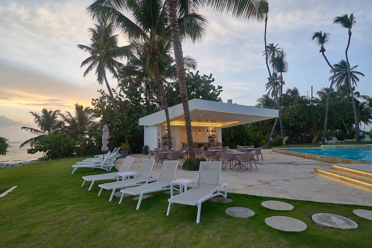 A spacious outdoor area is featured, with multiple lounge chairs arranged on green grass. A stylish structure with an overhanging roof provides shaded seating for dining. Lush palm trees surround the space, enhancing the tropical ambiance with a view of the ocean in the background.
