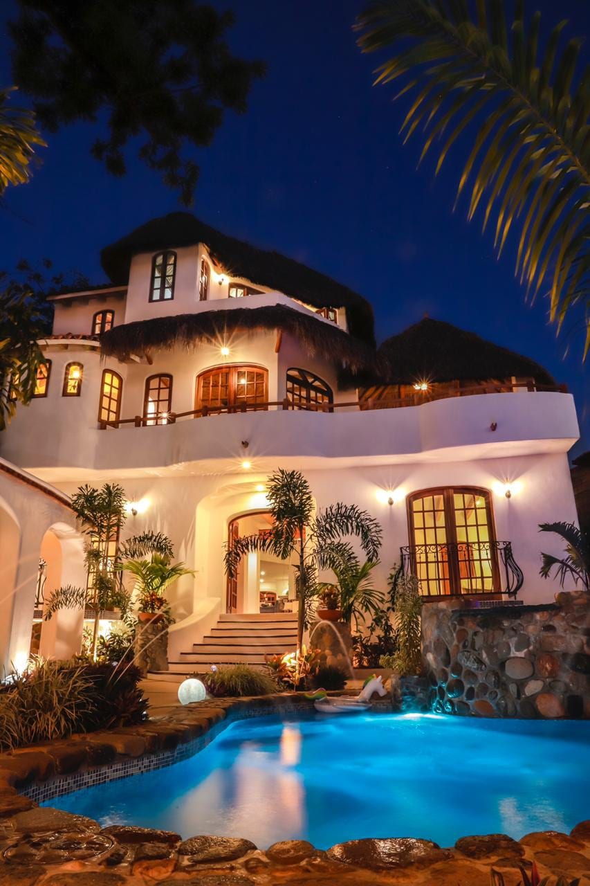 The exterior of the villa is showcased at night, with the warm glow of lights illuminating the structure. A stone pool is visible in the foreground, surrounded by tropical plants and staircases leading to the entrance, which features large arched windows.
