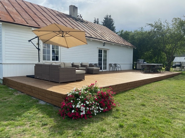 A spacious wooden terrace is highlighted, featuring comfortable seating under a large umbrella. A dining table is positioned to the side, with fresh flowers in bloom nearby. The white house exterior and natural greenery contribute to a relaxed outdoor setting.