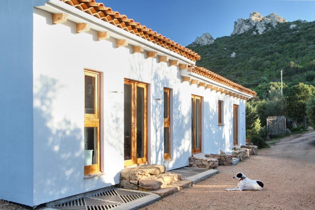 The exterior of the house is shown, featuring a white facade with wooden window frames. A stone pathway leads up to the entrance, with a dog resting nearby. The background displays lush greenery and rocky hills under a clear blue sky.