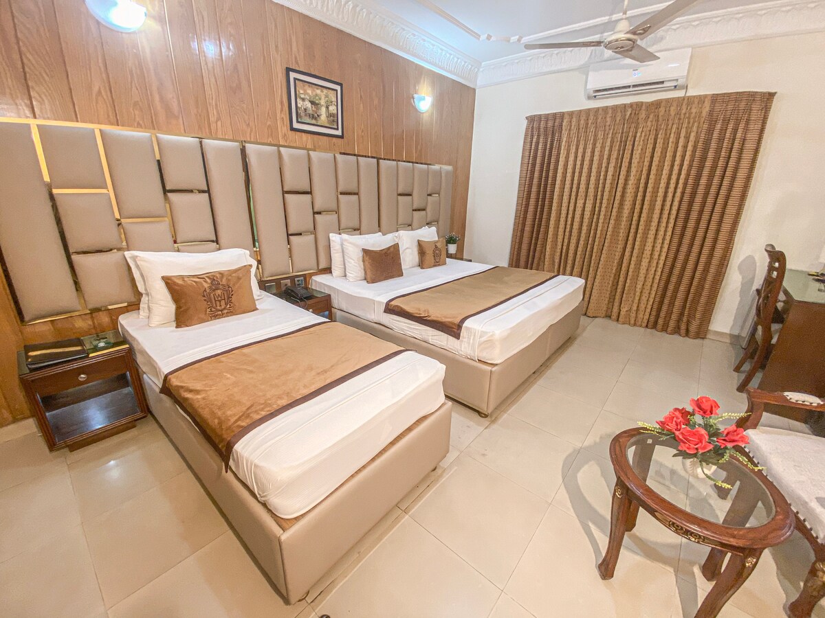 The guest room is equipped with two spacious beds adorned with warm brown and cream bedding. Elegant wall panels in light tones frame the area. A small sitting arrangement with a table and two chairs is present, while floor-to-ceiling curtains provide privacy.