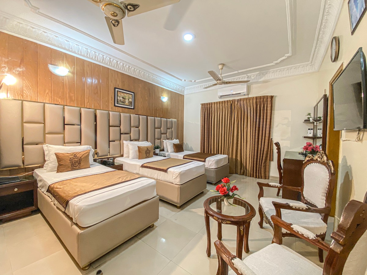 A spacious room is furnished with two large beds and a single bed, all with light-colored bedding. A small seating area features two elegant chairs and a round table. Warm wood paneling and softly lit lamps create a calm environment, while a large window is adorned with curtains.