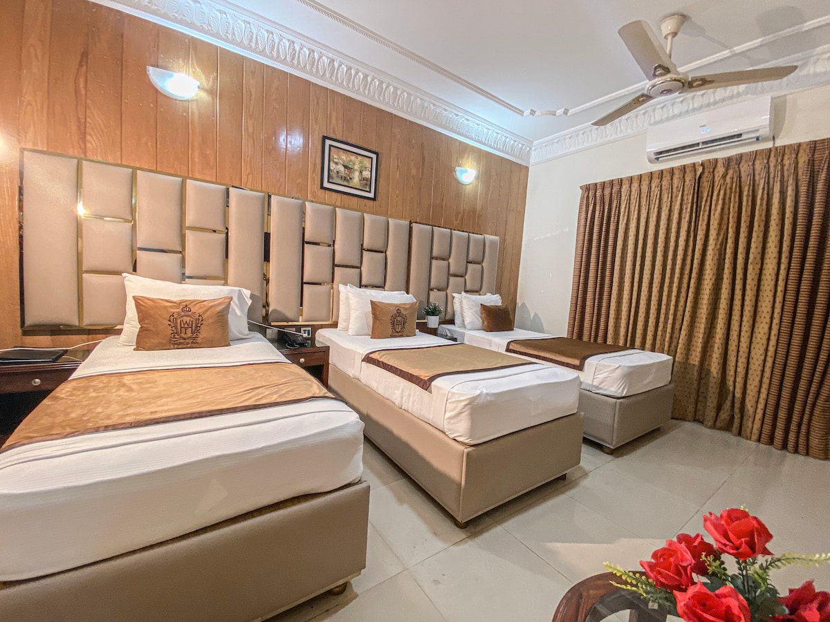 A well-appointed guest room features three spacious beds with tasteful linens. The wooden accent walls create a warm environment. A small table is placed next to the beds, with a decorative plant and red roses adding a touch of color. Soft lighting illuminates the space.