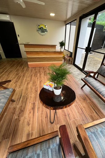 The reception area features a wooden check-in desk, complemented by light wooden flooring. Two wooden chairs with striped cushions are arranged around a circular table, adorned with a plant and a few magazines. Large glass doors provide natural light and an unobstructed view of the outdoor space.