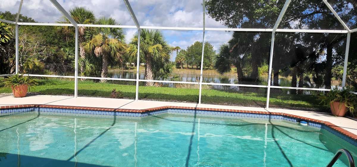 A screened-in pool area is shown, featuring clear waters and a tiled border. Lush palm trees and green foliage are visible in the background, while a serene lake is seen beyond the landscaping. The spacious area invites relaxation and enjoyment of the outdoor views.