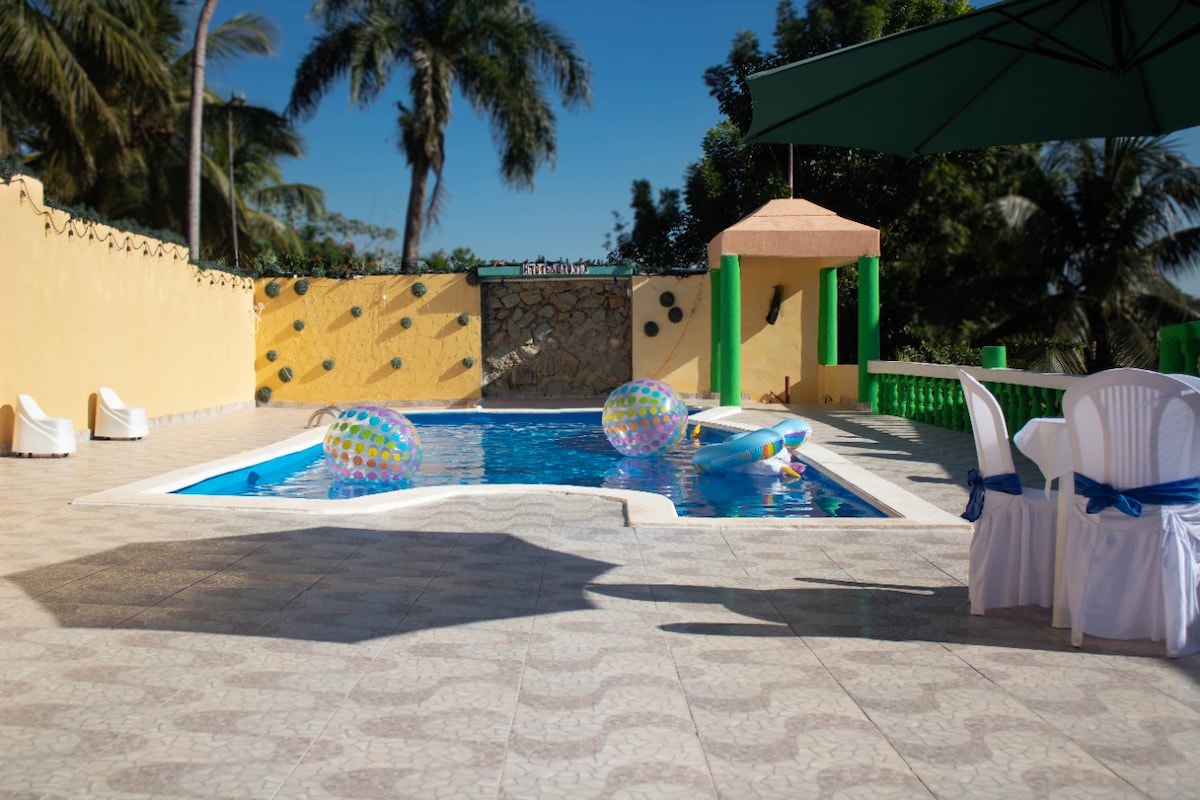A refreshing pool area is surrounded by vibrant yellow and green walls, featuring a clear blue water pool. Colorful inflatable floats rest on the water's surface. White chairs, adorned with blue sashes, are arranged nearby, offering a comfortable seating option in a sunny outdoor space.