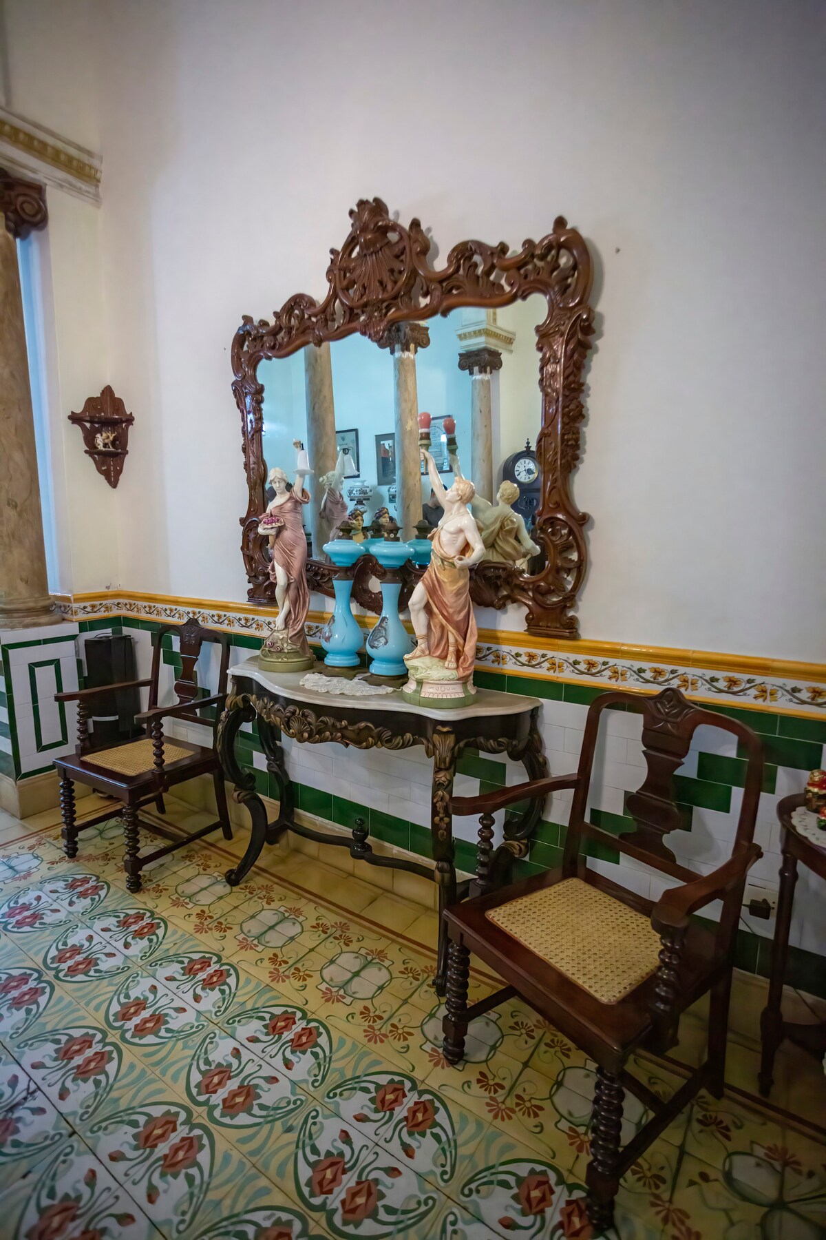 A vintage console table is highlighted against a decorated wall, featuring an ornate wooden mirror above it. The table is complemented by ceramic figures and vases. Two wooden chairs are positioned nearby on a colorful patterned tiled floor, characteristic of the colonial style.