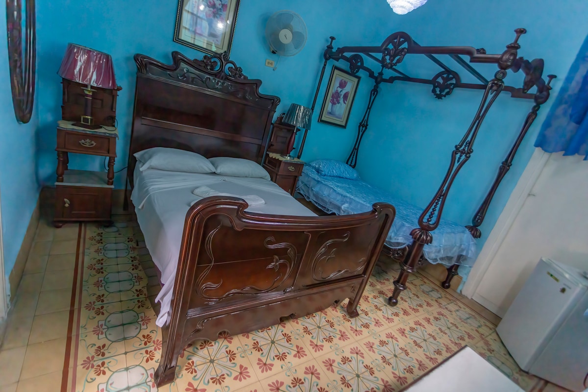 The bedroom features a wooden bed frame with a spacious mattress and an additional bed beside it. Blue walls create a serene backdrop, complemented by decorative blue curtains. Traditional tile flooring adds character, while a small nightstand and a fan enhance the room's functionality.