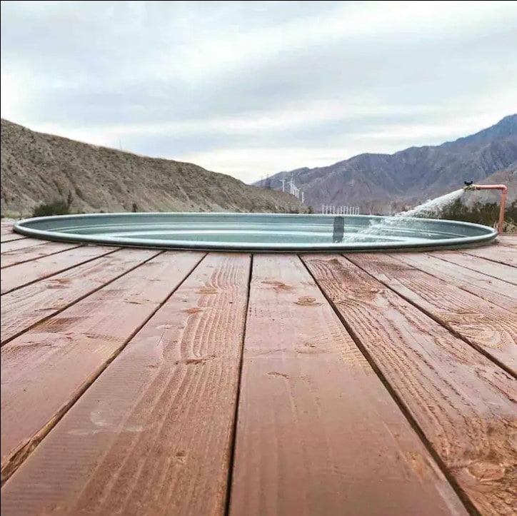A circular cowboy tub is positioned on a wooden deck, offering a tranquil escape. Surrounding the tub are sweeping views of the mountains in the distance, with a gentle stream of water flowing into it. The expansive natural landscape provides a serene backdrop.