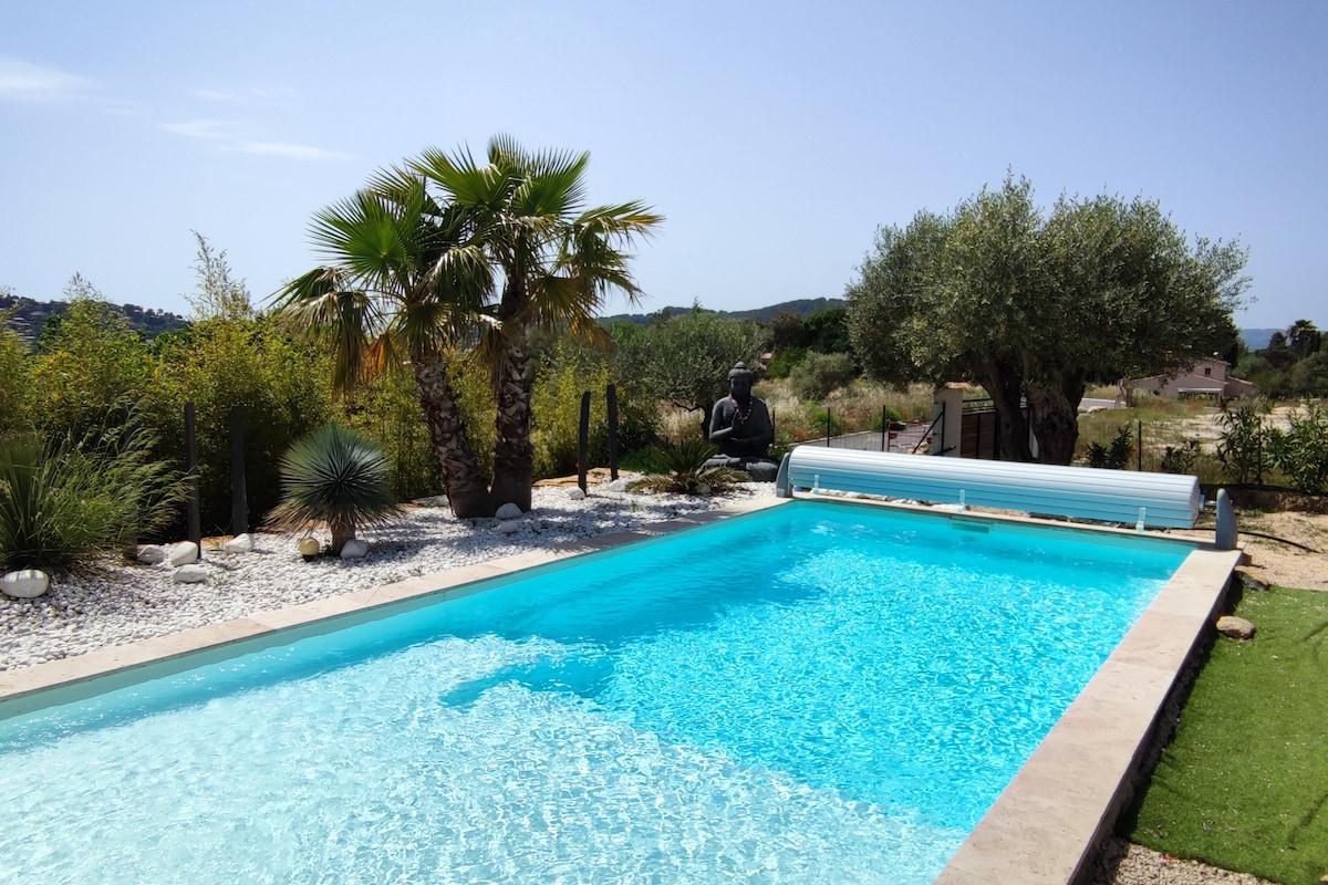 A rectangular salt-water pool is framed by natural vegetation, including palm trees and olive trees. The pool features a light blue water surface that reflects the sunlight, and a safety cover is rolled up beside the pool. A serene landscape surrounds the area.