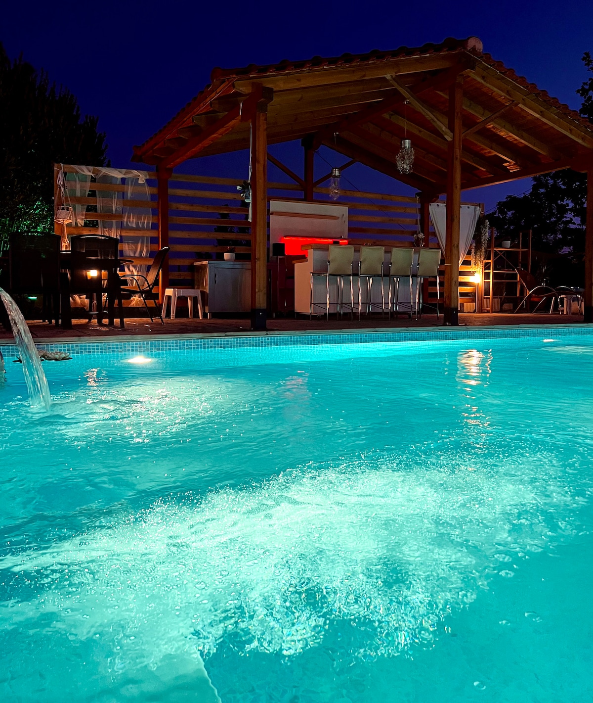 An illuminated pool reflects shades of turquoise under the night sky, with a cozy outdoor bar area visible in the background. The structure features a wooden roof and seating, creating a welcoming space for relaxation and enjoyment.