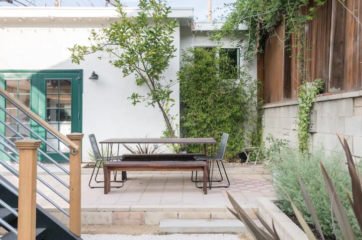 An outdoor dining area features a wooden table surrounded by metal chairs on a patterned stone patio. Lush greenery and a small tree provide natural elements, while a wood fence and a stairway add structure to the space.