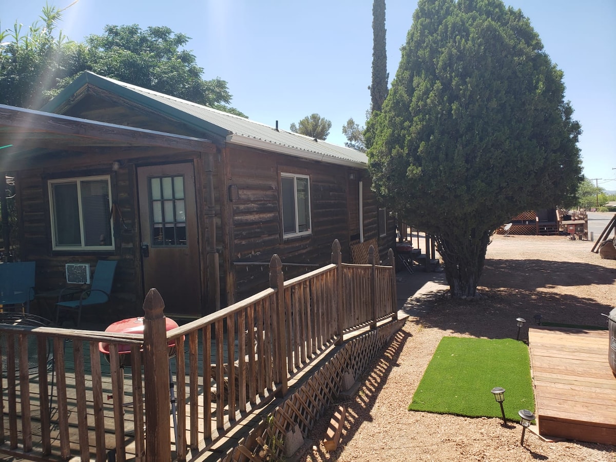 A rustic cabin made of logs is featured, set within a landscaped area that includes shrubs and a manicured patch of grass. Wooden steps lead to the entrance, while outdoor seating is arranged nearby. The cabin's windows are framed with green accents, merging with the surroundings.