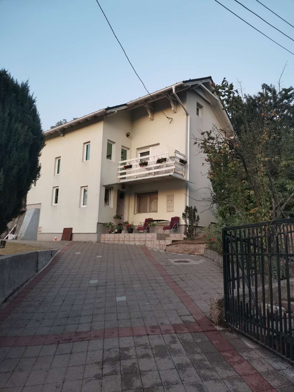 A three-story house is set within a landscaped yard, surrounded by trees and greenery. The front porch features a small balcony with white railings. A paved path leads to a fenced entrance, providing access to the property.