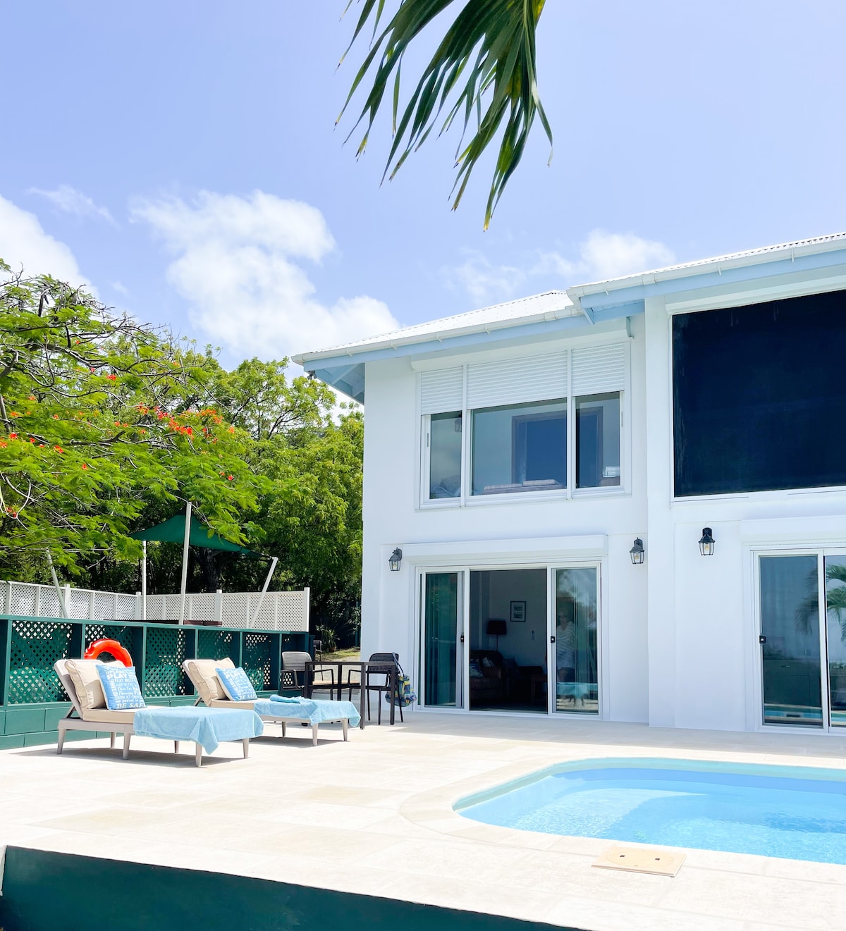 A modern villa presents a private pool with a sunbed area, surrounded by lush greenery. Large glass doors provide access to the interior, allowing natural light to fill the space. A shaded area with comfortable seating is visible in the background, promoting relaxation.
