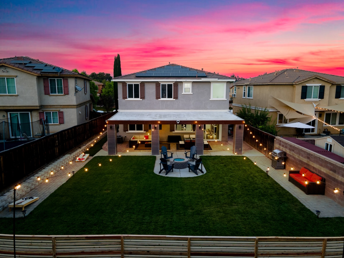 A spacious backyard is depicted at dusk, featuring a green lawn, a fire pit surrounded by chairs, and ambient lighting illuminating the area. A covered patio with outdoor seating is present, along with cornhole boards set near the fence. The two-story house is visible in the background.