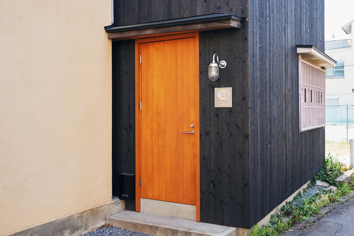 An entrance door is featured, crafted from polished wood and framed by a simple metal lantern. The surrounding wall is a mix of dark wood and beige, creating a modern aesthetic. A small outdoor area with gravel and greenery is visible beside the entrance.